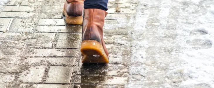 Walk on wet melted ice pavement. Back view on the feet of a man walking along the icy pavement. Pair of shoe on icy road in winter. Abstract empty blank winter weather background
